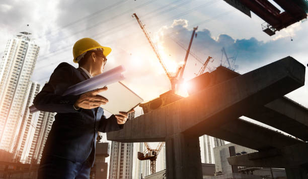 Construction engineer manager supervising progress of BTS Station Construction Project and tablet with blueprint in hands. Cranes in sunset background. Industrial technology 4.0