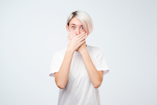 Close up of pretty young shocked woman with short colored hair covering mouth with her hands standing against gray background. I will keep your secret forever.