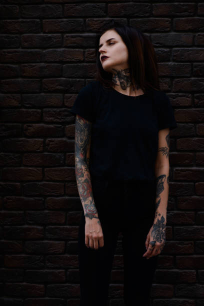 黒い唇、入れ墨とピアスのレンガの壁の上に立っている黒い服を着て美しいスリムなブルネットの少女 - tattoo t shirt brick wall portrait ストックフォトと画像