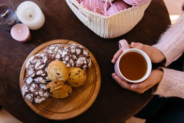 Cup of black tea and plate of fresh homemade cookies on wooden table with woolen coils of thread. Woman hands holding hot aromatic tea. Morning breakfast, warm and cozy