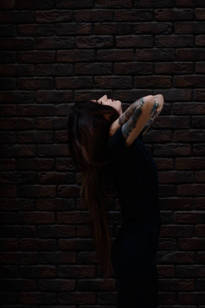 黒い唇、入れ墨とピアスのレンガの壁の上に立っている黒い服を着て美しいスリムなブルネットの少女 - tattoo t shirt brick wall portrait ストックフォトと画像