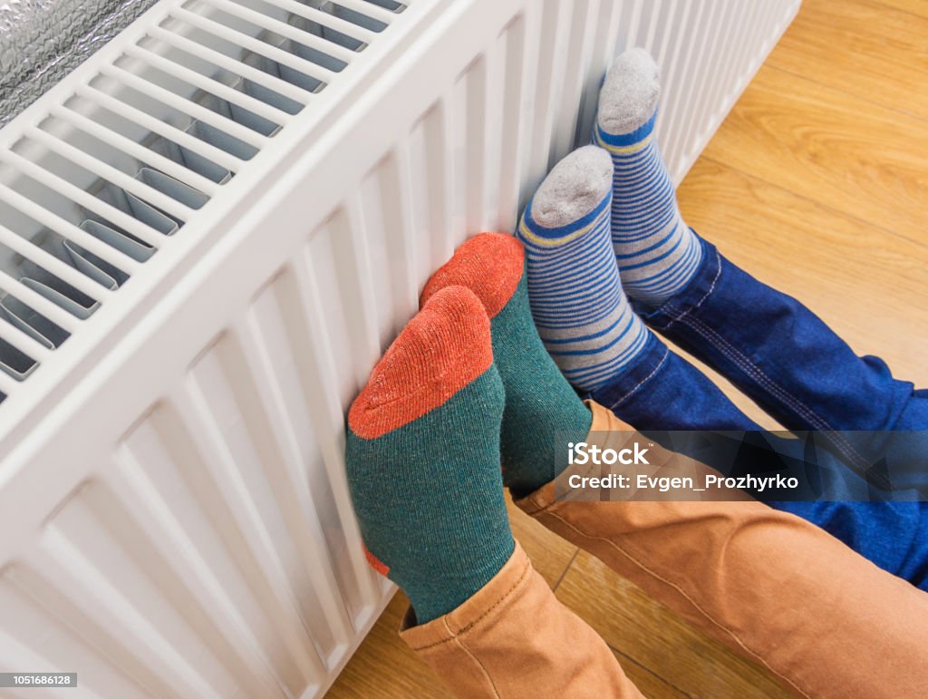Woman and child wearing colorful pair of woolly socks warming cold feet in front of heating radiator in winter time. Electric or gas heater at home. Woman and child wearing colorful pair of woolly socks warming cold feet in front of heating radiator in winter time. Electric or gas heater at home. Part of body, selective focus. Heat - Temperature Stock Photo