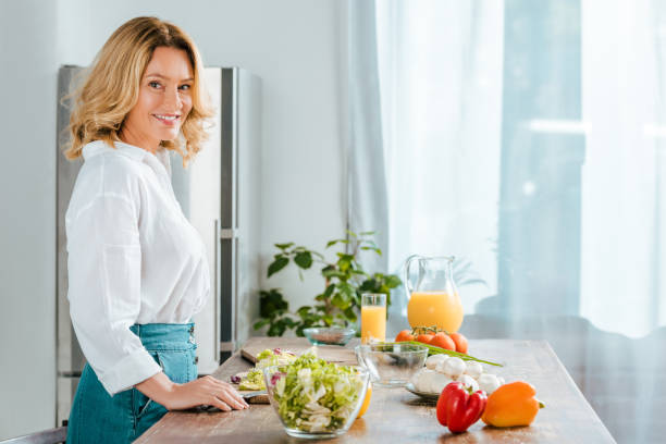 vista lateral de la mujer adulta feliz mirando a cámara haciendo la ensalada en la cocina - healthy eating food and drink raw leek fotografías e imágenes de stock