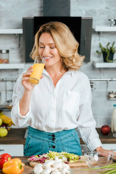 hermosa mujer adulta bebiendo jugo de naranja durante la cocción en la cocina - healthy eating food and drink raw leek fotografías e imágenes de stock