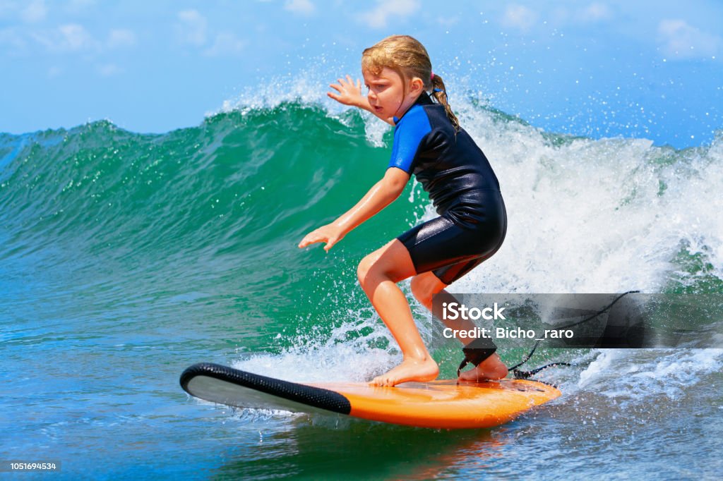 Young surfer rides on surfboard with fun on sea waves Happy baby girl - young surfer ride on surfboard with fun on sea waves. Active family lifestyle, kids outdoor water sport lessons and swimming activity in surf camp. Summer vacation with child. Surfing Stock Photo