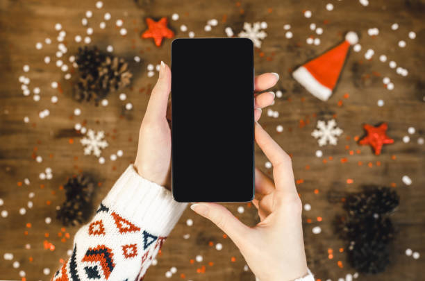 クリスマス ギフト雪と雪で木製の背景に oled ディスプレイとスマートな携帯電話を保持している女性の手。新年あけましておめでとうございます、携帯電話上のサンタの帽子、フラット ト� - retina display ストックフォトと画像