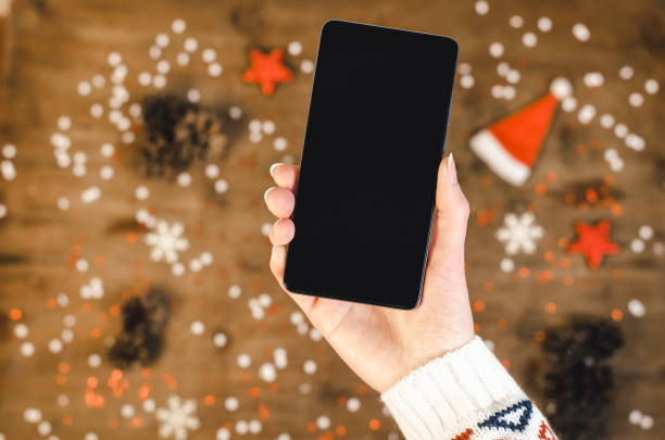 クリスマス ギフト雪と雪で木製の背景に oled ディスプレイとスマートな携帯電話を保持している女性の手。新年あけましておめでとうございます、携帯電話上のサンタの帽子、フラット ト� - retina display ストックフォトと画像