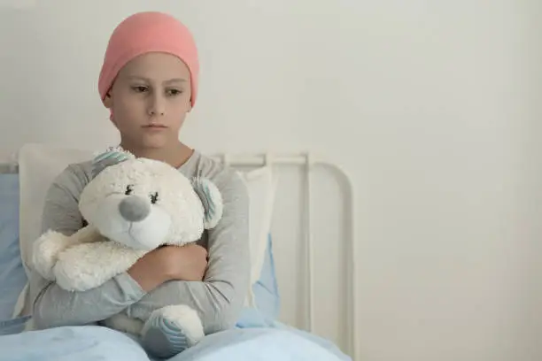 Weak girl with cancer wearing pink headscarf and hugging teddy bear next to copy space