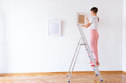 Woman on the ladder fitting picture frames on the white wall