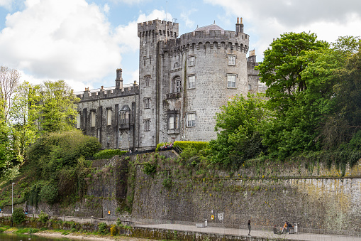 KILKENNY, LEINSTER, IRELAND - MAY 12, 2018: medieval Kilkenny castle round tower close-up from the bridge crossing river Nore between lush vegetation, with people walking leisurely on the promenade.