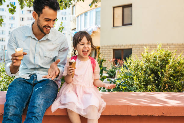 Image of happy cute little girl sitting with dad on the city street and eating ice-cream outdoor. Fun girl kid and father have fun and playing outside. Good relationship between dad and daughter stock photo