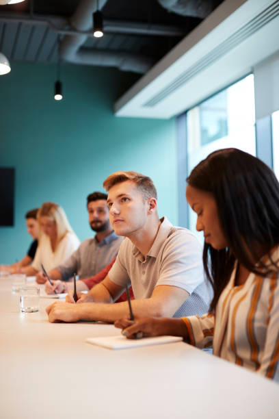 istock Group Of Young Candidates Sitting At Boardroom Table Making Notes At Presentation At Business Graduate Recruitment Assessment Day 1051777578
