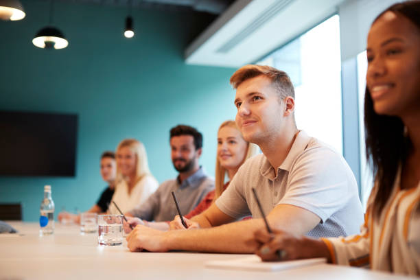 Group Of Young Candidates Sitting At Boardroom Table Listening To Presentation At Business Graduate Recruitment Assessment Day Group Of Young Candidates Sitting At Boardroom Table Listening To Presentation At Business Graduate Recruitment Assessment Day trainee stock pictures, royalty-free photos & images