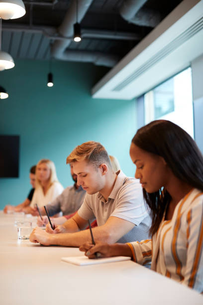 istock Group Of Young Candidates Sitting At Boardroom Table Making Notes At Presentation At Business Graduate Recruitment Assessment Day 1051777586
