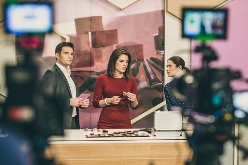 Photo of two blurred cameras filming three people trying make-up for a television infomercial.