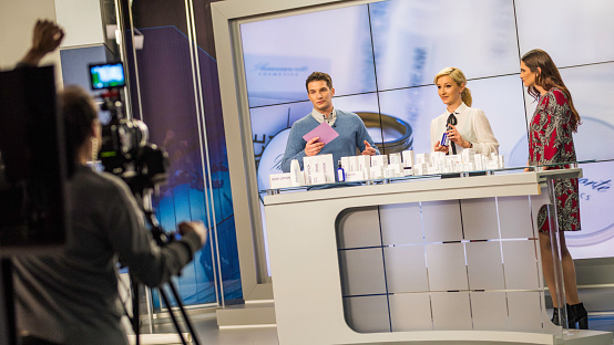 A man and two women standing next to a counter full of cosmetic products being filmed by a camera while talking about them.