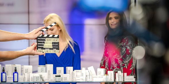 Photo of a pair of hands holding a clapboard saying 'body cream' and two women at a counter before the start of filming of a body cream infomercial.