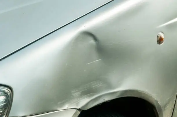 Dented sheet metal on the side of the silver car damaged in crash accident on the parking lot or in the traffic