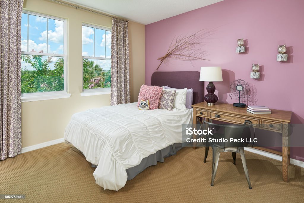 A Pink and Tan Little Kids Bedroom with an Owl Theme A ruffled style white comforter sits under pink, white, and purple pillows. There's a grey underskirt for the full size bed above the tan, pimpled, carpet floor. A deep purple fluffy headboard sits at the head of the bed. Next to the bed, there is a thin wood desk with three drawers and a metallic, wired, metal chair. On top of the desk are three books, a metal flower for holding jewelry, and a lava lamp looking, deep purple lamp with a white lampshade. A bundle of sticks and three owl figurines on top of round wood pieces hang on a light purple wall. The opposite wall is tan with two windows, looking out to a foliage of trees and a blue sky dotted with clouds. The curtains patterned cream and purple matches some of the pillows on the bed. Bedroom Stock Photo