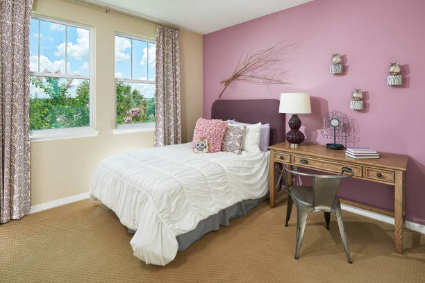 A Pink and Tan Little Kids Bedroom with an Owl Theme A ruffled style white comforter sits under pink, white, and purple pillows. There's a grey underskirt for the full size bed above the tan, pimpled, carpet floor. A deep purple fluffy headboard sits at the head of the bed. Next to the bed, there is a thin wood desk with three drawers and a metallic, wired, metal chair. On top of the desk are three books, a metal flower for holding jewelry, and a lava lamp looking, deep purple lamp with a white lampshade. A bundle of sticks and three owl figurines on top of round wood pieces hang on a light purple wall. The opposite wall is tan with two windows, looking out to a foliage of trees and a blue sky dotted with clouds. The curtains patterned cream and purple matches some of the pillows on the bed. tidy room stock pictures, royalty-free photos & images
