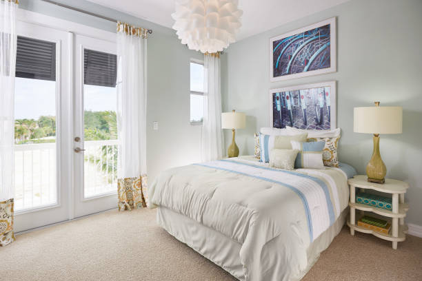 A Blue, White, and Off-White, Bedroom with a Bicycle Theme in a Model Home A large bed sits next to a double door leading out to a small balcony overlooking a sandy seascape. The wall is a blue gray color, and the doors and windows are white. The curtains are see through with fringe orange and blue patterning. On the ceiling is an interesting chandelier of white overlapping cups, creating a flower effect. On the wall above the bed hangs two photos of back, black bicycle tires, blue tire guards, and red, oval reflectors. Below, on the bed, sits two long white pillows, two pillows matching the drapery, and three pillows matching the white, light blue, and tan bed spread. On the four leaf clover, sandy white end tables sits a yellow lamp with a twist in its neck and a white lamp shade. Three books of various sizes and colors are on the end table in shot. The floor is a brown carpet. tidy room stock pictures, royalty-free photos & images
