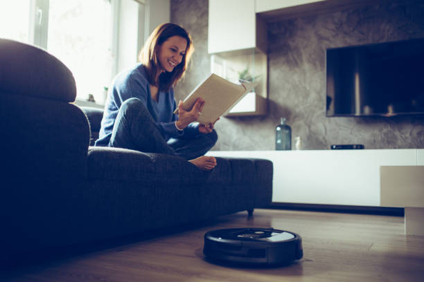 istock Woman reading book while robotic vacuum cleaner cleaning house 1052175816