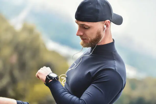 Photo of Muscular bearded athlete checking burned calories on smart watch after good workout session on city park.