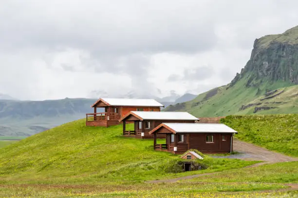 Three rustic wooden cabins in Vik, Iceland with cloudy, overcast grey stormy sky and cliff during summer, camping hotel accommodation