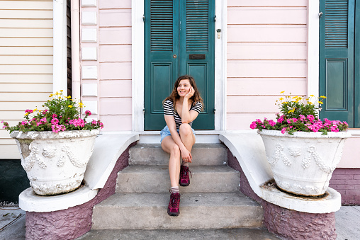 Young hipster millennial woman sitting smiling happy on stairs steps, front porch in New Orleans by colorful pink door architecture with yellow flowers in spring pot