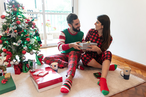 Beautiful young couple exchanging christmas presents while wearing christmas pajamas both looking very happy