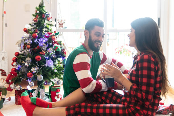 junges paar, lachen und reden und genießen sie einen kaffee tragen ihre weihnachts-pyjamas und weiter an den weihnachtsbaum sitzen - communication discussion coffee christmas stock-fotos und bilder