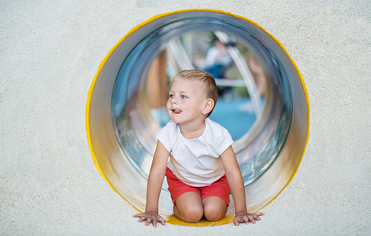 A little boy enjoys the underground multicolored tube on the playground on a sunny summer day.