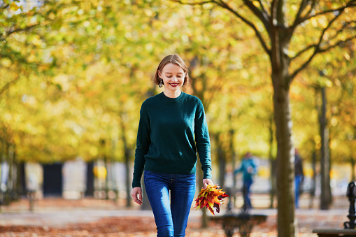 Beautiful young woman with bunch of colorful autumn leaves walking in park on a fall day. Luxembourg garden, Paris, France