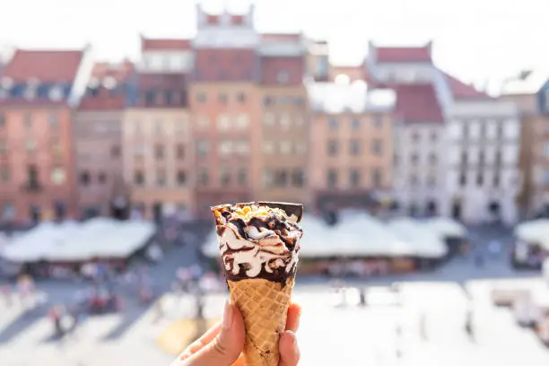 Woman hand holding one white vanilla chocolate ice cream gelato cone with background of Warsaw, Poland old market square historic buildings in town city during hot sunny summer day