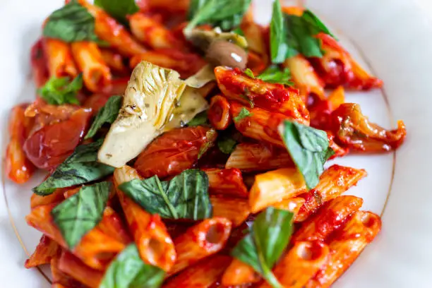 Flat top table, closeup of fresh vibrant red tomato sauce penne pasta corn gluten free noodles on plate in marinara with chopped basil greens, artichokes in restaurant on white plate