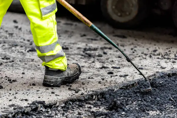 Closeup of construction worker with neon yellow green pants clothing, boots, raking hot black wet tar and asphalt, bitumen in city site in London