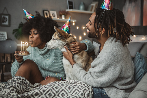 Young couple on bed enjoy in small celebration. Theay are celebrating birthday with dog. Blowing candles on cake and wearing party hats.