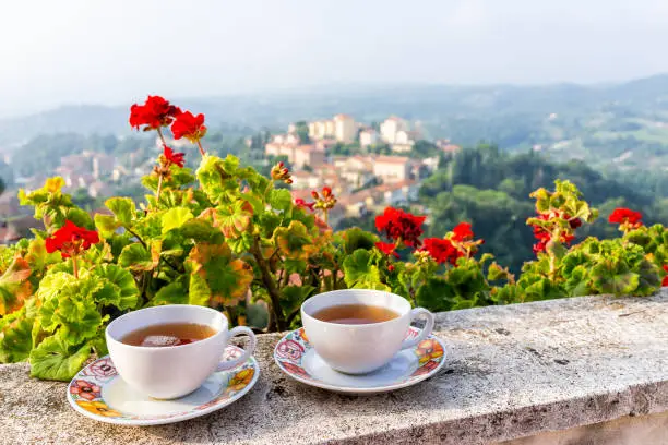 Two tea cups on balcony terrace by red geranium flowers outside in Italy with mountain view of Chiusi cityscape, Umbria near Tuscany at sunset
