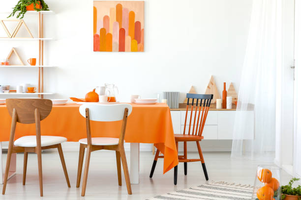 istock Real photo of kitchen table covered with orange tablecloth and white dishes. Artwork on the wall and shelf in the corner 1052675652