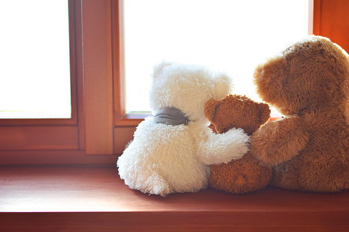 Three toy bears - mother father and child sitting on brown window sill hugging each other and looking out of window against sunlight back view. Love, family and parenthood concept.