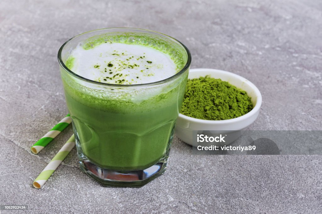 Organic green matcha tea latte in glass and powder in white bowl Organic green matcha tea latte in glass and powder in white bowl with cocktail straw on gray concrete background Matcha Tea Stock Photo