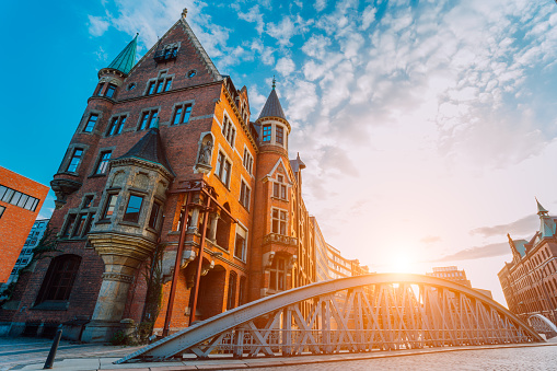 Metal arch bridge and old red bricks building in the Speicherstadt warehouse district of Hamburg HafenCity with sunburst light during sunset golden hour and white clouds against blue sky above