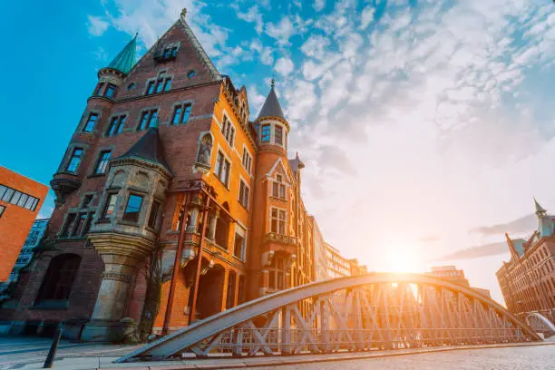 Metal arch bridge and old red bricks building in the Speicherstadt warehouse district of Hamburg HafenCity with sunburst light during sunset golden hour and white clouds against blue sky above.