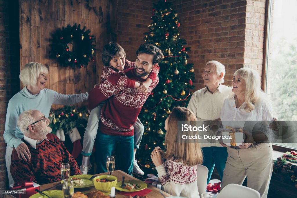 Noel morning large gathering, couple meeting, fairy decoration december tradition. Grey-haired grandparents, grandchildren, relatives, brother, son, at house feast lunch table, dad piggybacking girl Christmas Stock Photo