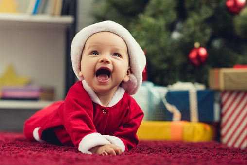 close-up portrait of laughing little baby in santa suit lying on red carpet in front of christmas tree and gifts