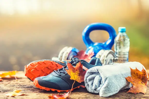 Pair of blue sport shoes water and  dumbbells laid on a wooden board in a tree autumn alley with maple leaves -  accessories for run exercise or workout activity.