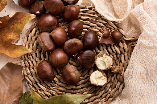 high angle view of some chestnuts out from a paper cone, on a rustic wicker mat, on a table next to some autumn leaves