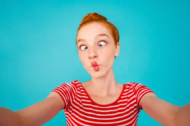 Photo of Self-portrait of crazy foolish playful pretty ecstatic cheerful red-haired girl with bun, grimacing, isolated on bright vivid blue background