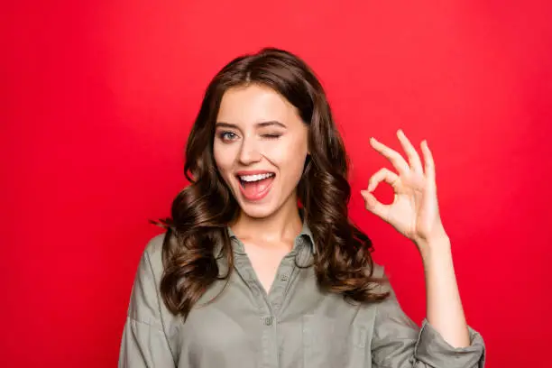 Close up portrait of sweet, gorgeous, nice, adorable, good-looking lady with modern curly hairstyle, casual shirt, show Ok sign and wink isolated on shine red background