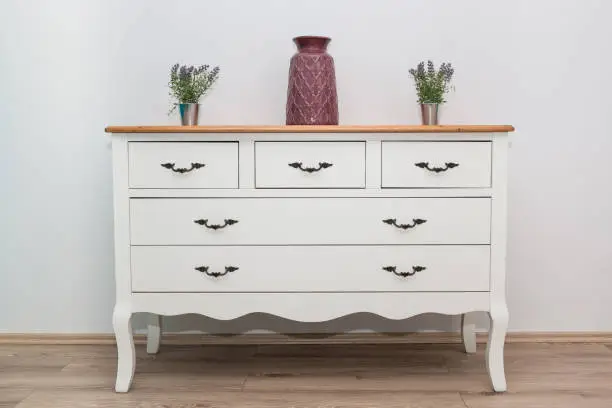 Photo of White wooden dresser with three vases and flowers on white wall background. Chest of drawers close up.